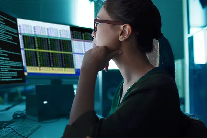 A woman in glasses focuses intently on dual computer screens displaying complex data in a dimly lit office. The atmosphere is analytical and concentrated.