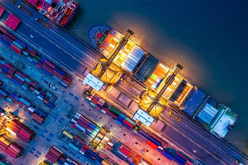 Aerial view of a busy shipping port at night. Illuminated cargo ship is docked, surrounded by colorful containers on the pier, creating a vibrant scene.