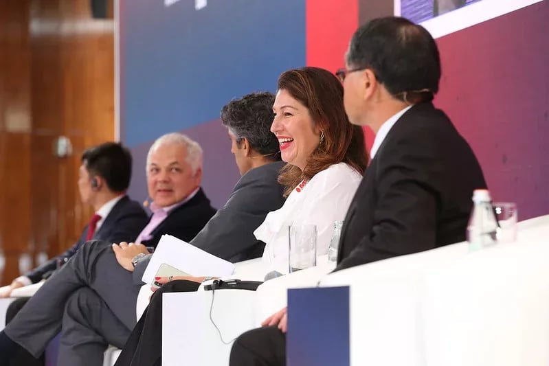 A panel discussion at a professional conference, featuring five diverse panelists seated on a stage. A woman in a white outfit is smiling and engaging in conversation, while the other panelists, dressed in formal business attire, listen attentively. The background features a modern conference setting with a red and blue-themed stage backdrop. Water bottles and microphones are placed in front of the panelists, indicating a structured discussion.