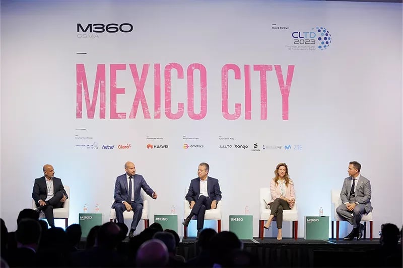A panel discussion at the M360 GSMA event in Mexico City. Five professionally dressed panelists, including four men and one woman, are seated on a stage engaging in conversation. One panelist in a blue suit is gesturing while speaking. The backdrop prominently features 'MEXICO CITY' in bold pink letters, along with event sponsor logos such as Telcel, Claro, Huawei, ZTE, and others. The audience is visible in the foreground, attentively listening to the discussion. The setting suggests a high-profile industry conference focused on digital transformation and the technology ecosystem.