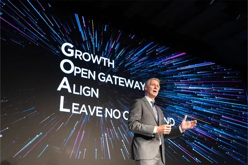 A professional speaker in a grey suit and tie is delivering a presentation on stage. He gestures with both hands while speaking. Behind him, a large digital screen displays the acronym 'GOAL' with its meaning: 'Growth, Open Gateway, Align, Leave No One Behind.' The background features a dynamic, futuristic design with streaks of blue and purple light radiating outward, creating an engaging and high-tech atmosphere.