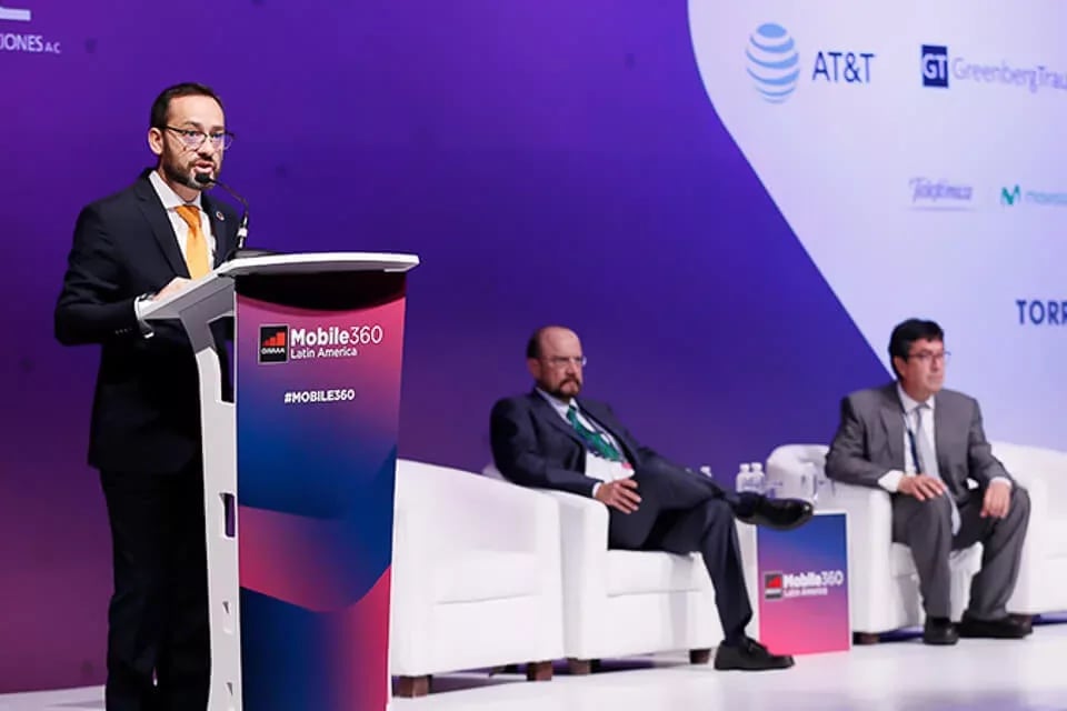 A professional conference setting at 'Mobile360 Latin America.' A man in a dark suit, white shirt, and yellow tie stands at a podium with a microphone, delivering a speech. Behind him, two panelists sit on white armchairs, dressed in business attire, attentively listening. The stage backdrop features a purple gradient design with sponsor logos, including 'AT&T' and 'Greenberg Traurig.' The event branding is prominently displayed on both the podium and the stage background.